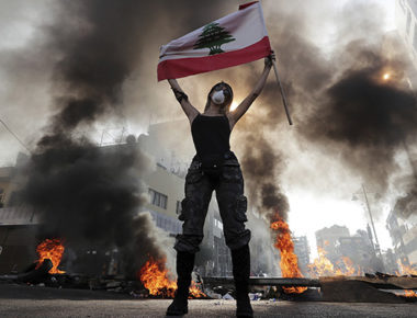 An anti-government protester holds a Lebanese flag while standing in front burning tires that block a road in the town of Jal el-Dib, north of Beirut, Lebanon, Wednesday, Nov. 13, 2019. Lebanese protesters blocked major highways with burning tires and roadblocks on Wednesday, saying they will remain in the streets despite the president's appeal for them to go home. (AP Photo/Hassan Ammar)